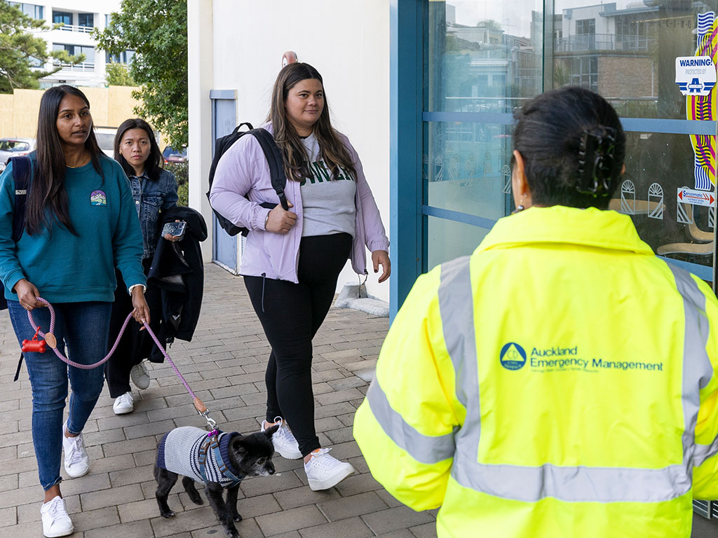 A group of people, one walking a dog, approaches a person in an Auckland Emergency Management jacket outside a building.